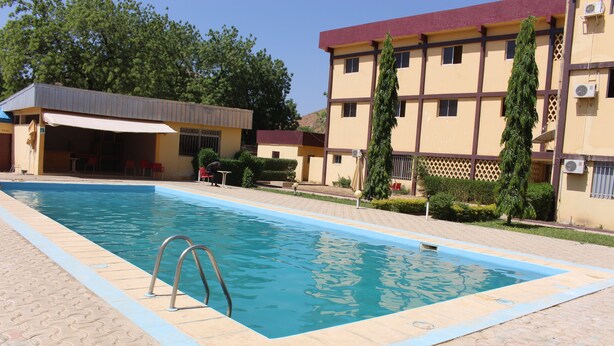 A swimming pool with a tiled deck, a covered seating area, and a building with multiple windows and air conditioning units. - Hotel Maroua Palace (Maroua)