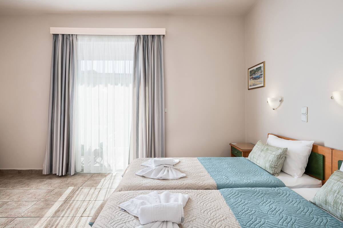 Quarto de hotel com duas camas, uma janela grande com cortinas, uma mesa de cabeceira e um candeeiro fixado na parede.