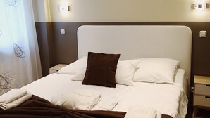 Una cama bien hecha con ropa de cama blanca y marrón, una almohada marrón y toallas dobladas. En la pared sobre la cama, hay un cuadro de un puente.