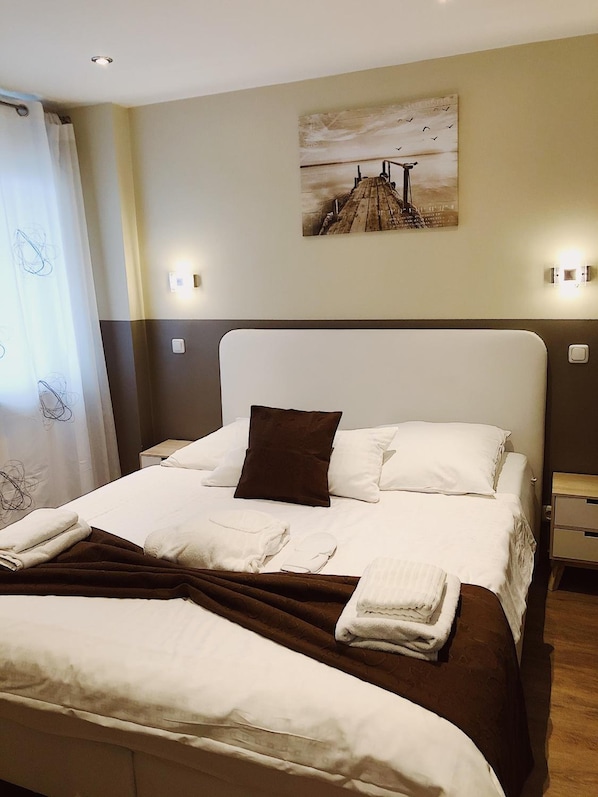 Una cama bien hecha con ropa de cama blanca y marrón, una almohada marrón y toallas dobladas. En la pared sobre la cama, hay un cuadro de un puente.