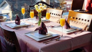 Una mesa de desayuno servida con platos, cubiertos, vasos de zumo de naranja y un jarrón con flores.