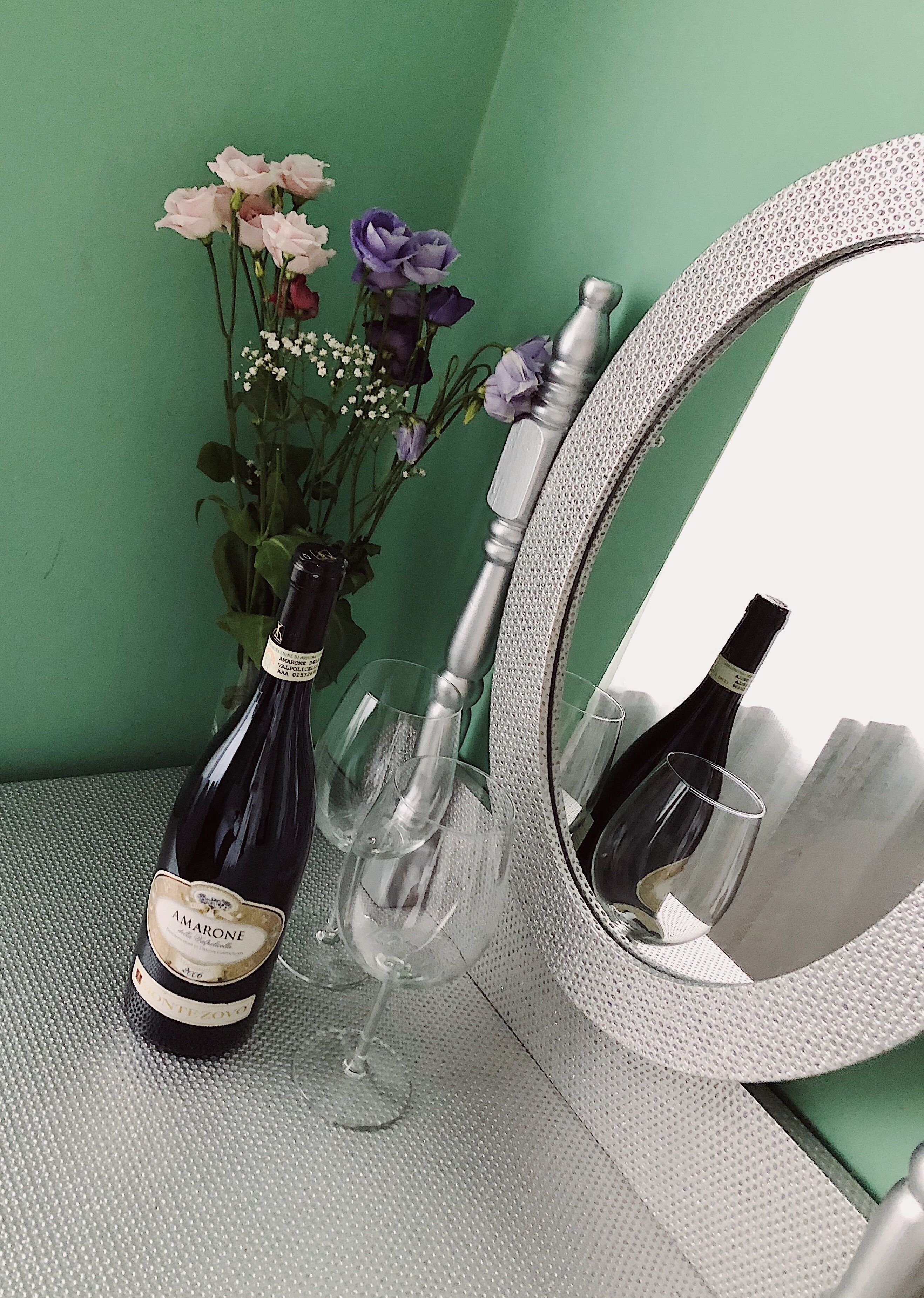 A bottle of Amarone wine, a wine glass, and a mirror with a textured frame on a textured surface.