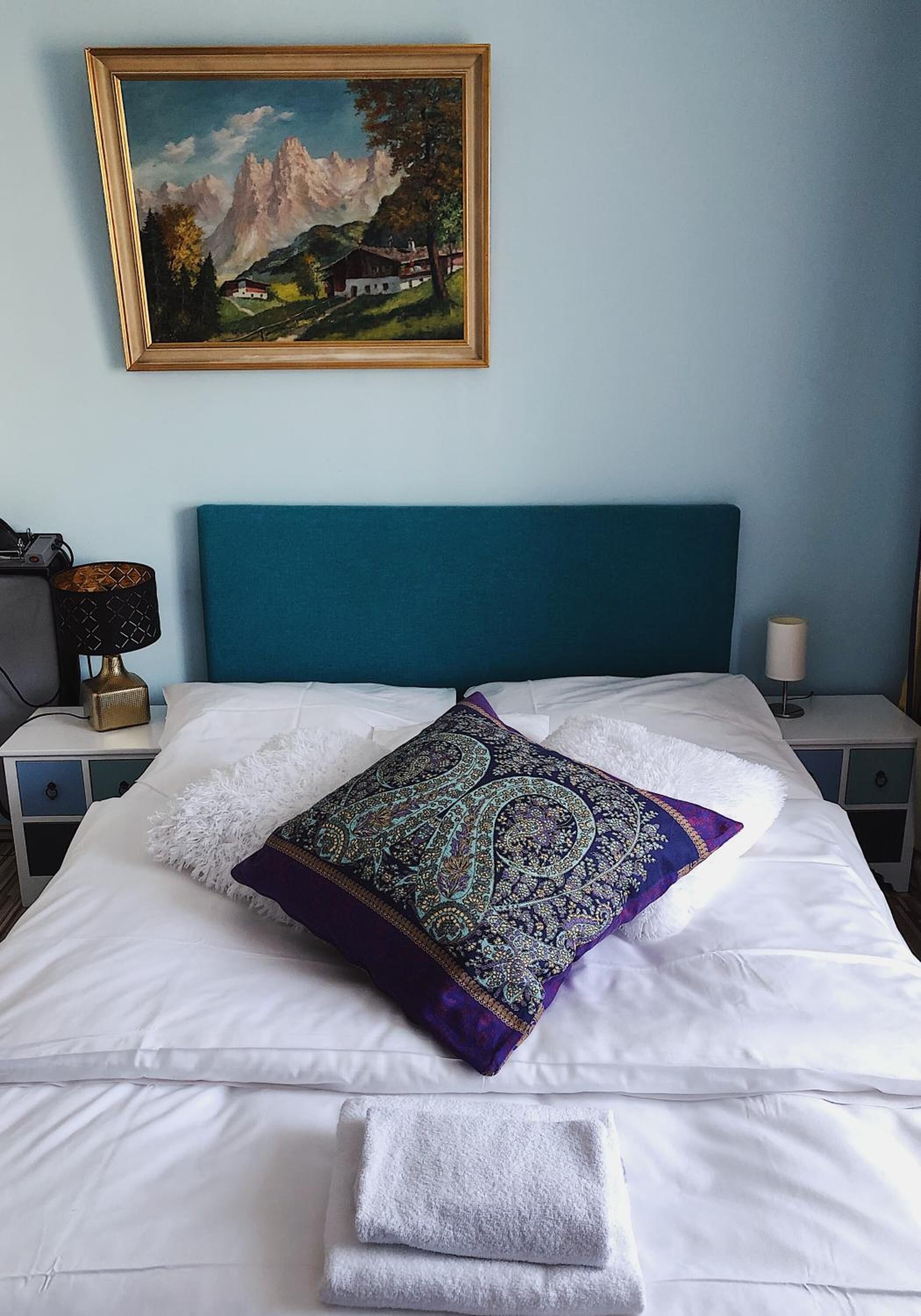 Un lit bien fait, avec des draps blancs, un coussin décoratif et une serviette pliée, posés sur une tête de lit bleue.