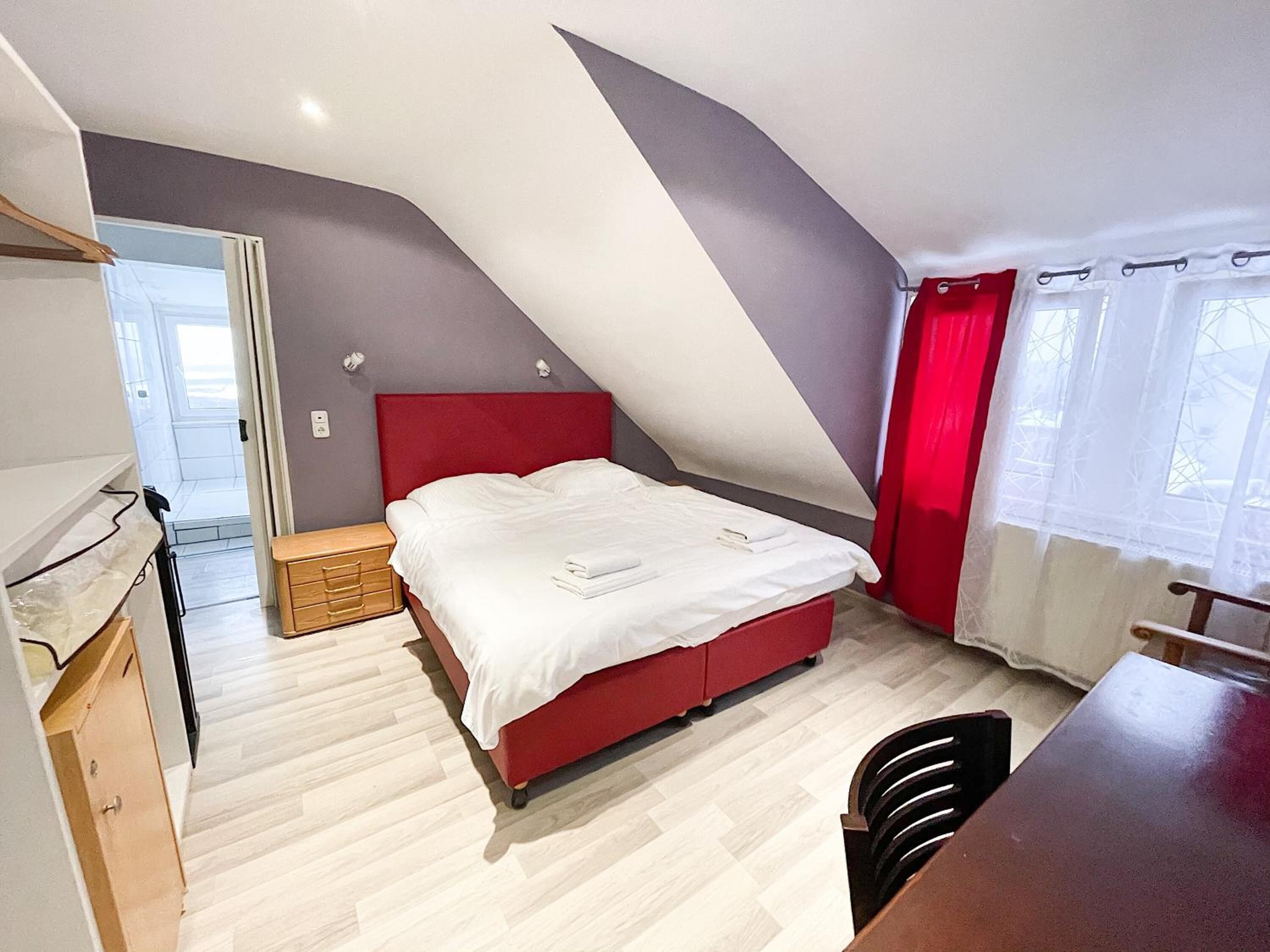 Un dormitorio con techo inclinado, cama roja, cómoda de madera y escritorio con silla.