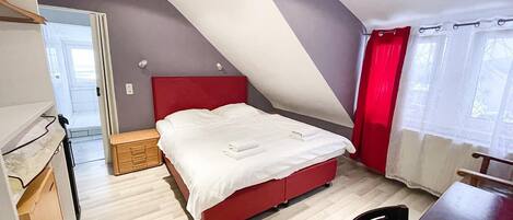 Un dormitorio con techo inclinado, cama roja, cómoda de madera y escritorio con silla.