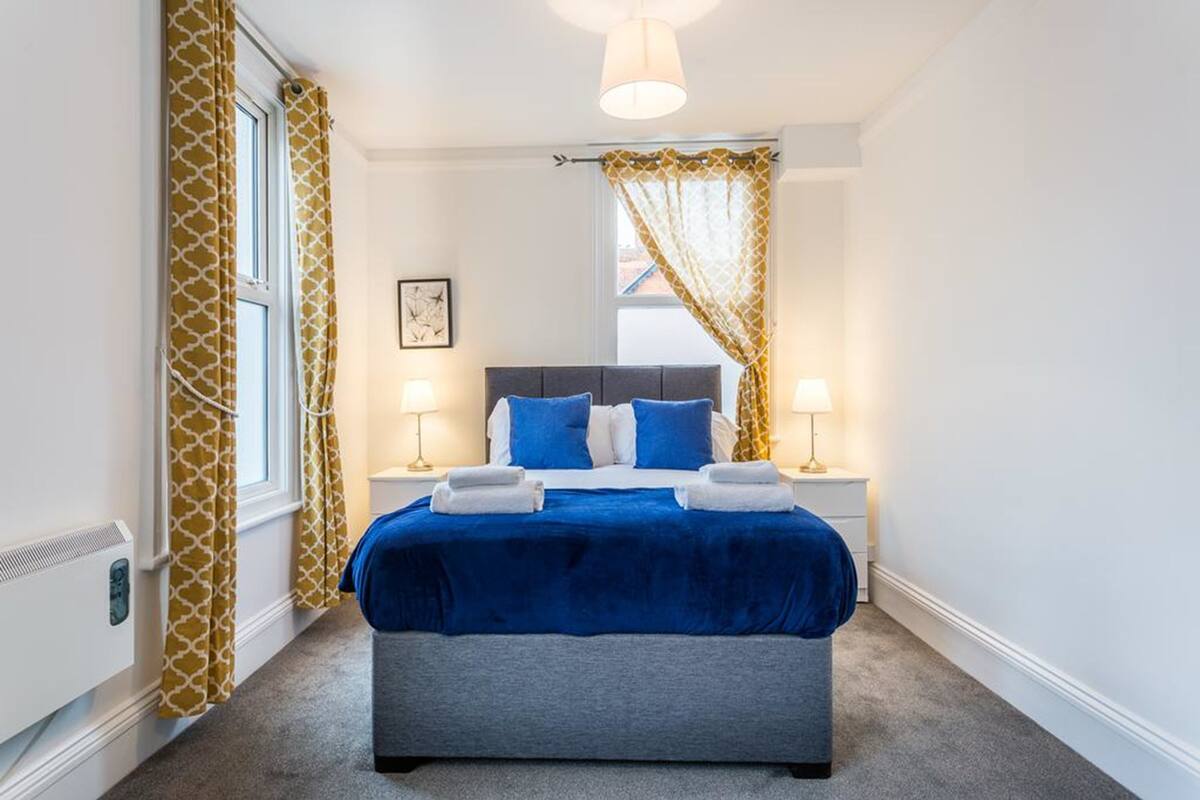 Een opgemaakt bed met blauw beddengoed, twee bedlampjes en een raam met patroon gordijnen.
