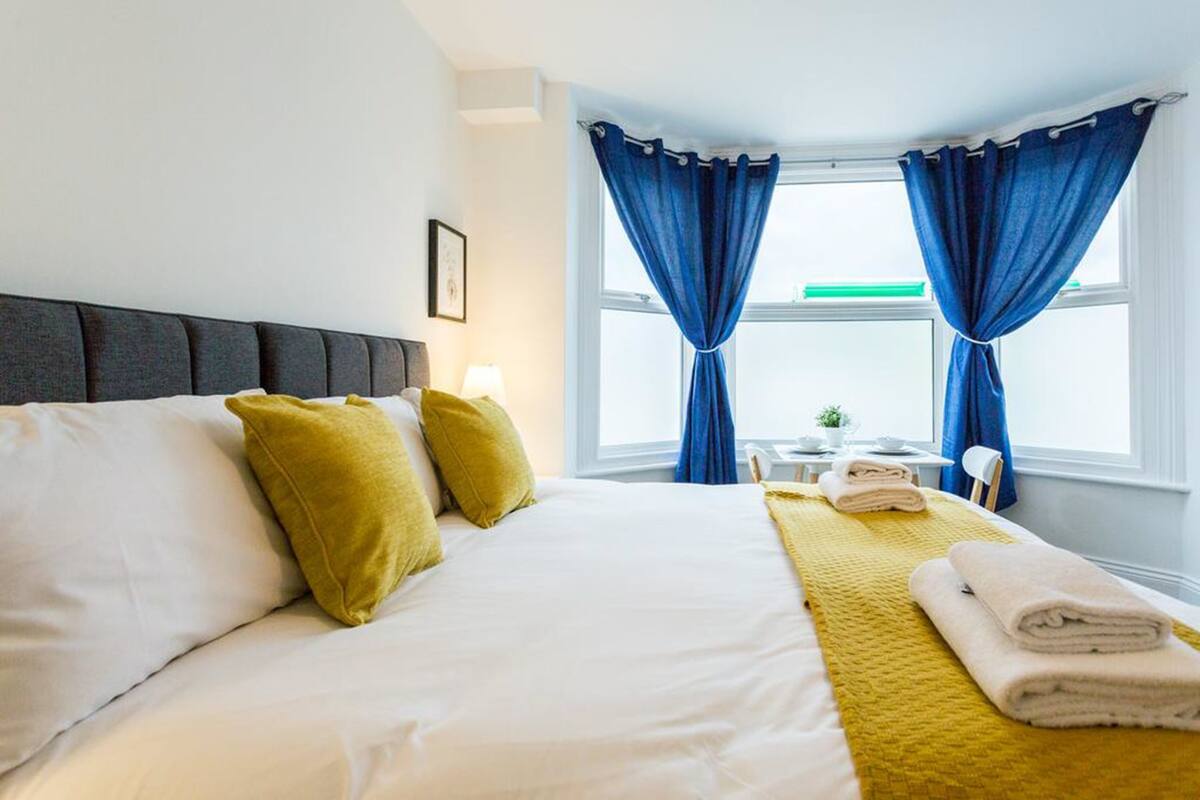 Een netjes opgemaakt bed met wit en geel beddengoed, een hoofdbord en een gele plaid.