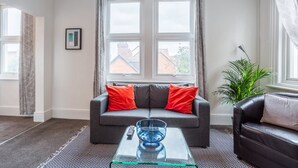 Ein modernes Wohnzimmer mit grauem Sofa, roten Kissen, einem Glastisch und einem großen Fenster mit Vorhängen.