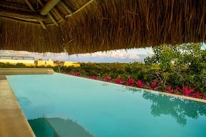 A swimming pool with a thatched roof overhead, surrounded by lush greenery and a clear sky. - Azul Tulum by GuruHotel (Tulum)