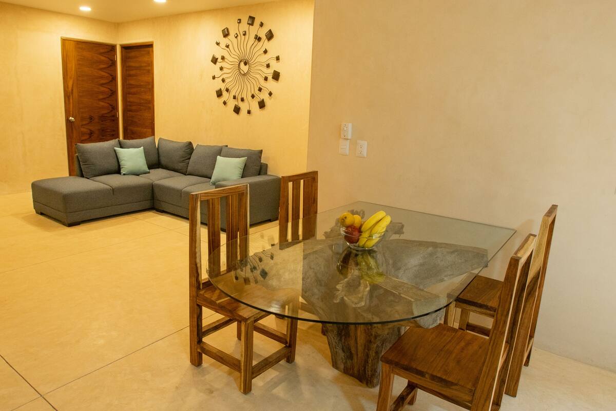 A dining area with a glass table and wooden chairs, a gray sofa with cushions, and a decorative wall piece.