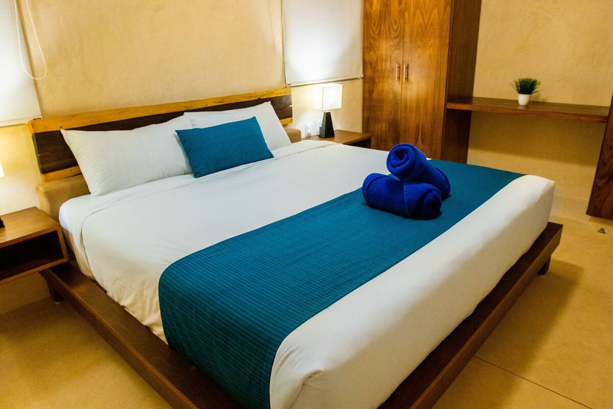 A neatly made bed with a teal bedspread and white linens, a wooden headboard, a nightstand with a lamp, and a small potted plant on a shelf.