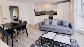 A modern living room with a grey sofa, a dining table with chairs, and a kitchen area with white cabinets and appliances. - Roomspace Apartments -Kew Bridge Court (London)