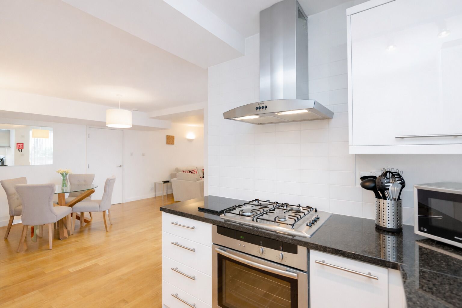 Une cuisine moderne équipée d’une cuisinière à gaz, d’un four et d’une hotte en acier inoxydable. On y trouve un coin repas avec une table et des chaises en verre, ainsi qu’une petite cuisine avec un four à micro-ondes et des ustensiles.