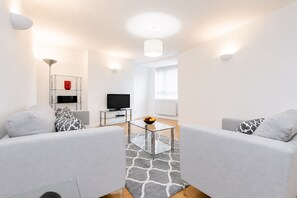 A modern living room with a sofa, a glass coffee table, a TV, and a red vase on a shelf. - Roomspace Apartments -Kew Bridge Court (London)