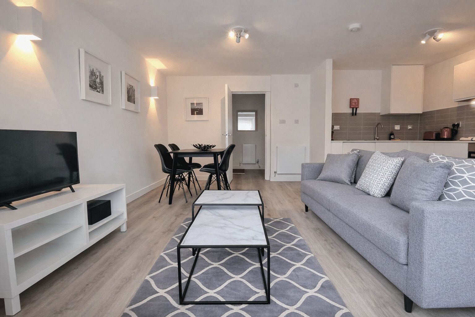 A modern living room with a grey sofa, a TV on a white stand, a dining area with a table and chairs, and a kitchen in the background.