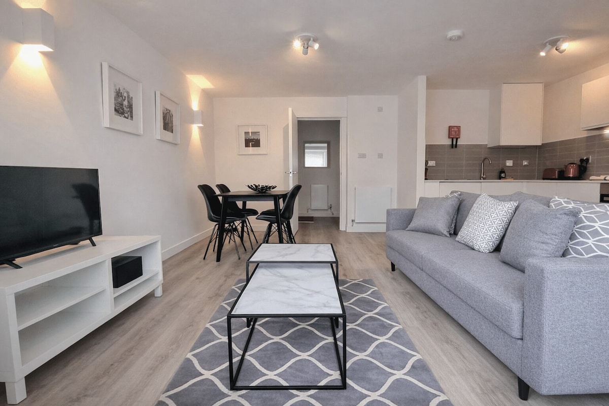 A modern living room with a grey sofa, a TV on a white stand, a dining area with a table and chairs, and a kitchen in the background.