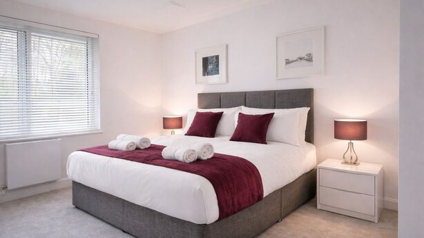 A modern bedroom with a grey upholstered bed, white linens, and a red throw blanket. There are two bedside tables with lamps, a window with blinds, and two framed pictures on the wall. - Roomspace Apartments -Kew Bridge Court (London)
