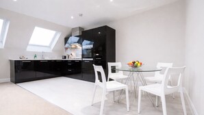 A modern kitchen with black cabinets, a white countertop, and a glass dining table with white chairs. - Roomspace Apartments -Kinnaird Court (Esher)