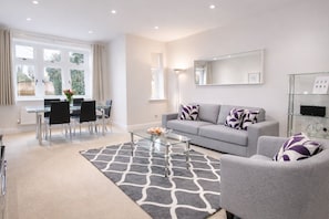 Uma sala de estar moderna com um sofá cinzento, uma mesa de centro de vidro e um espaço de refeições com uma mesa e cadeiras de vidro.