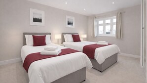 A hotel room with two beds, each with white linens and red pillows, a window with curtains, and two framed pictures on the wall. - Roomspace Apartments -Kinnaird Court (Esher)
