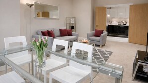 A modern living room with a glass dining table, white chairs, a grey sofa with red cushions, and a kitchen in the background. - Roomspace Apartments -Kinnaird Court (Esher)