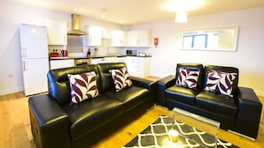 A modern living room with two black leather sofas, a glass coffee table, and a kitchen in the background. - Roomspace Apartments -Kirk Court (Dorking)