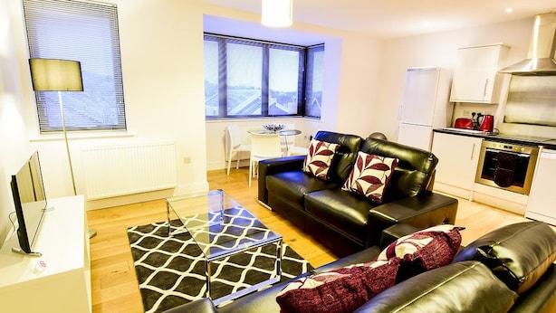 A modern living room with a black leather sofa, a glass coffee table, and a patterned rug. There is a kitchen area with a stove and cabinets. - Roomspace Apartments -Kirk Court (Dorking)