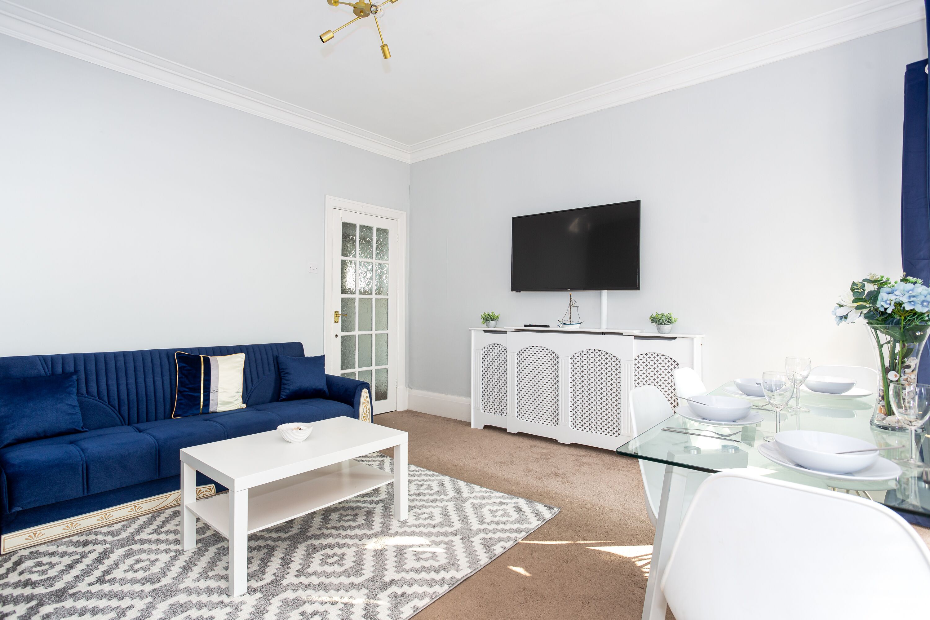 A modern living room with a blue sofa, white coffee table, and a glass dining table set for four.
