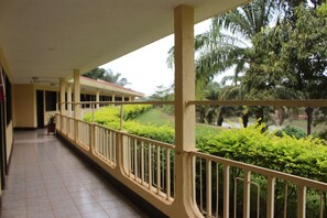 A covered corridor with a tiled floor, a railing, and a view of green bushes and trees. - Hotel Mansa (Bertoua)