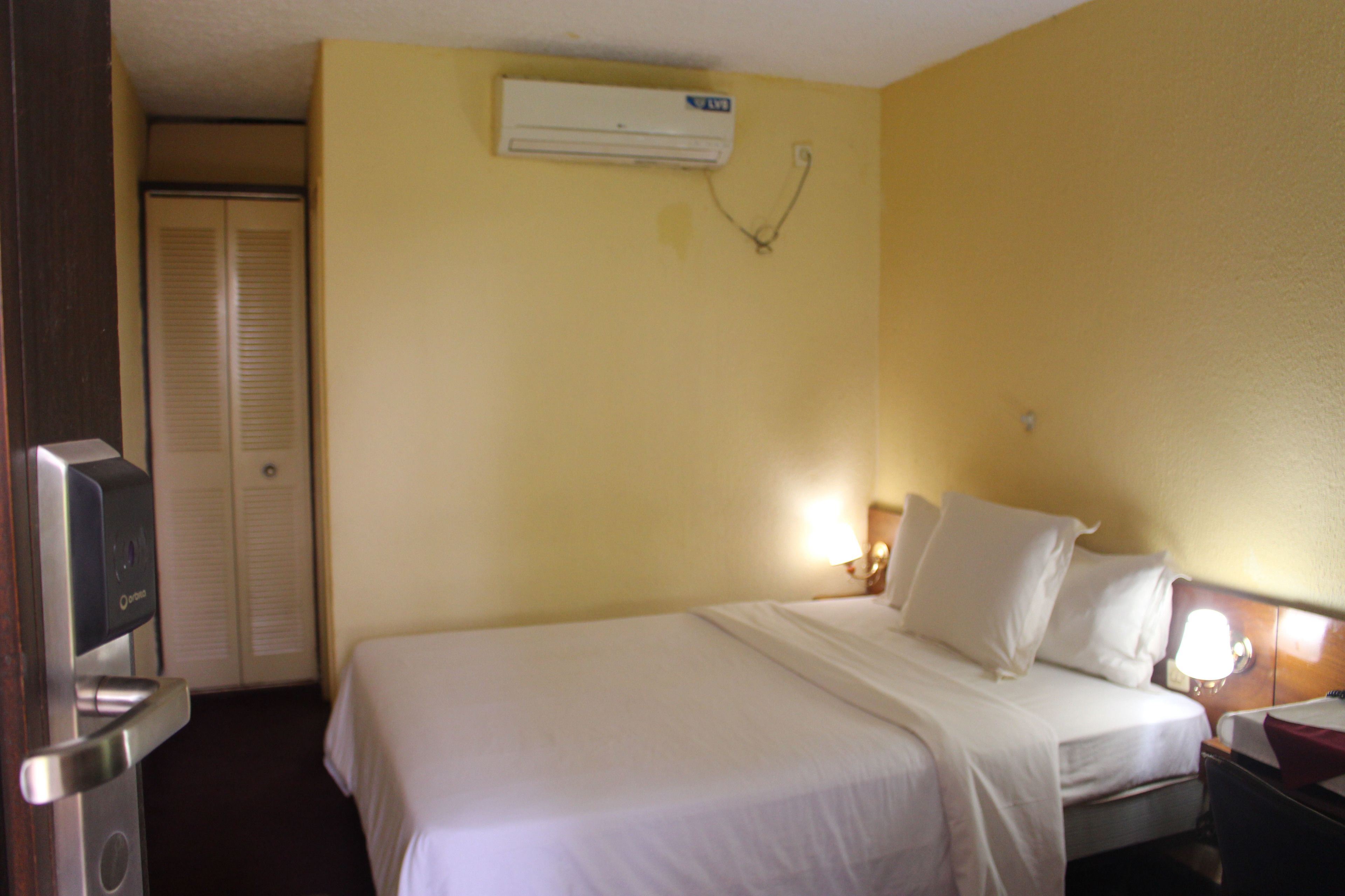 A hotel room with a bed, a bedside table, a lamp, an air conditioning unit, and a door with a mirror.