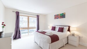 Quarto de hotel com uma cama grande, uma mesa de cabeceira com abajur, uma janela com cortinas e um quadro na parede.