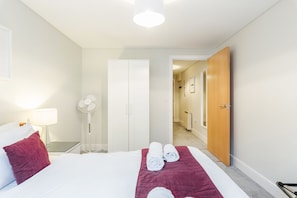 A hotel room with a bed, a bedside table with a lamp, a fan, and a door leading to a hallway.