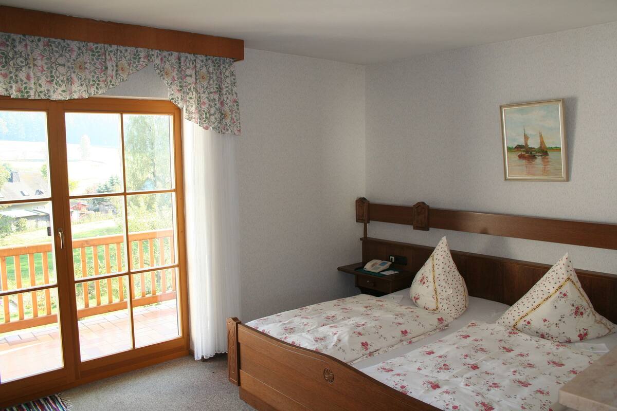 Un dormitorio con cama de madera, ropa de cama floral, una mesita de noche, un cuadro y un ventanal corredero a un balcón.