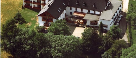 An aerial view of a large, multi-story building with a sloped roof, surrounded by dense trees and a grassy field.