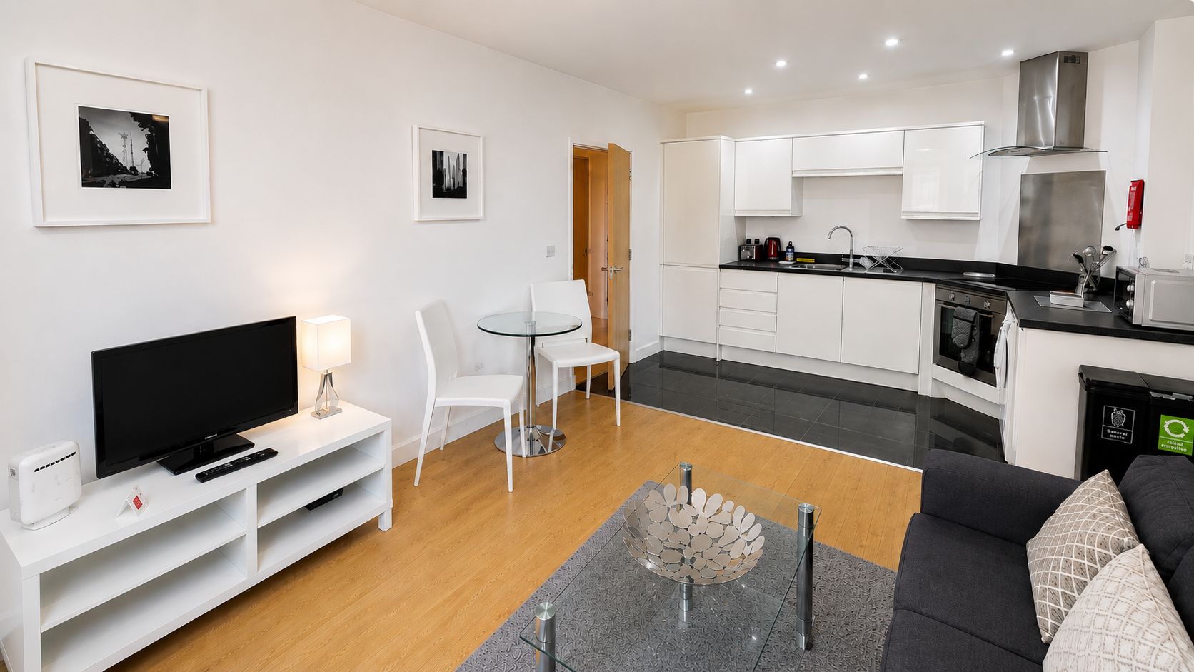A modern living room with a flat-screen TV, white furniture, a glass coffee table, and a grey rug.