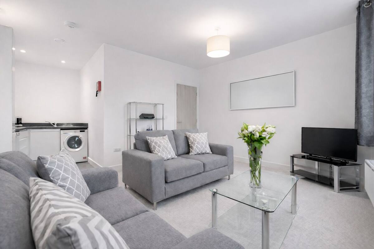 A modern living room with a grey sofa, a glass coffee table, a flat-screen TV, and a washing machine in the background.