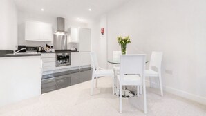 A modern kitchen with white cabinetry, a stainless steel range hood, and a dining area with a glass table and white chairs. - Roomspace Apartments -Nouvelle House (Sutton)
