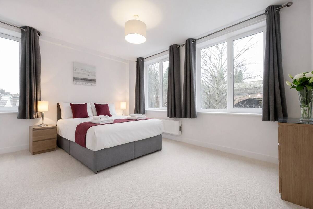 A modern bedroom with a large bed, bedside tables, a radiator, and a view of the outside through large windows.