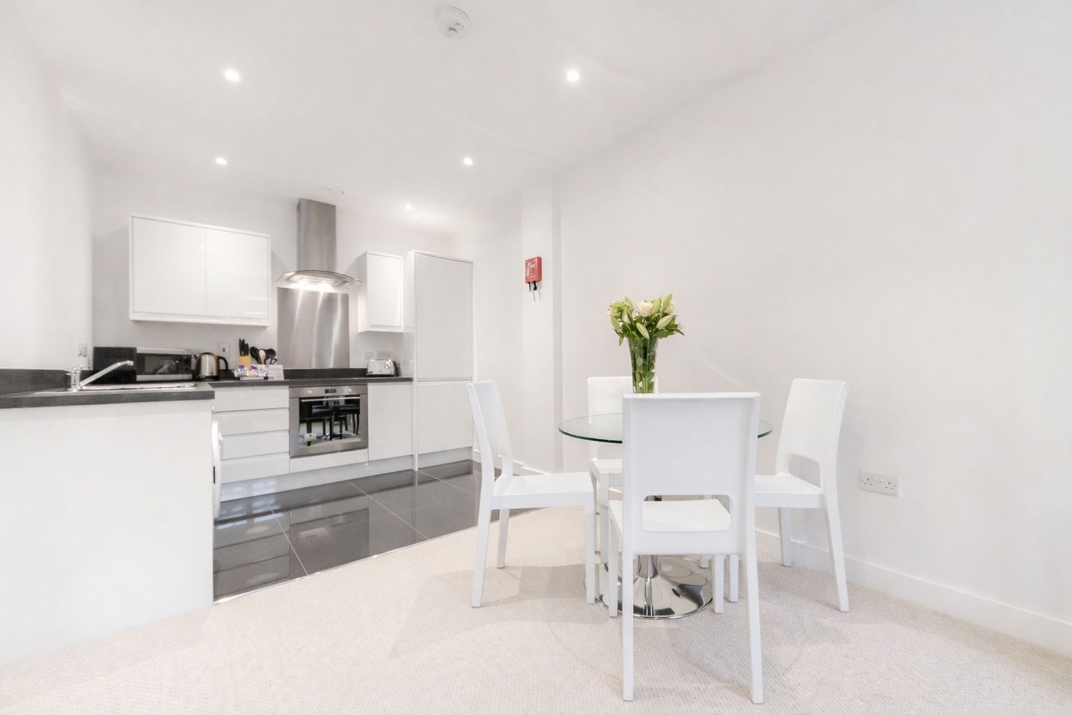 A modern kitchen with white cabinetry, a stainless steel range hood, and a dining area with a glass table and white chairs.