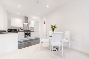 Une cuisine moderne avec des meubles blancs, une hotte en acier inoxydable et un coin repas comprenant une table en verre et des chaises blanches.