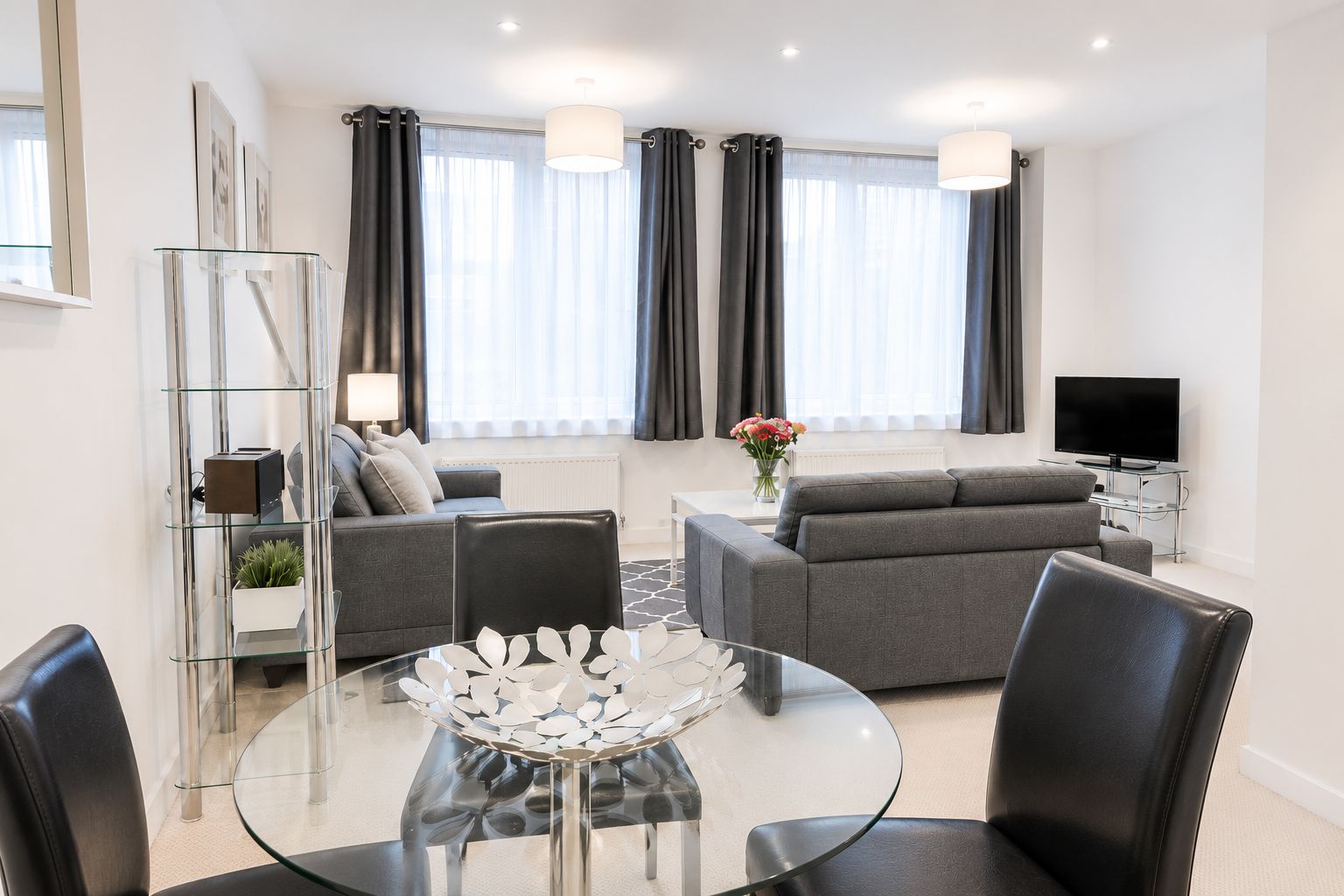 A modern living room with a glass coffee table, grey sofas, and a flat-screen TV.