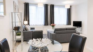 A modern living room with a glass coffee table, grey sofas, and a flat-screen TV.
