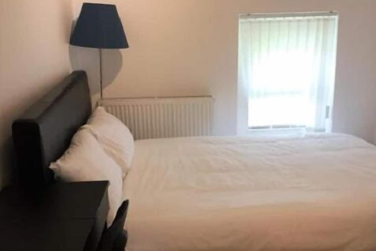 Een slaapkamer met een bed, een bureau, een stoel, een lamp en een raam met gordijnen.