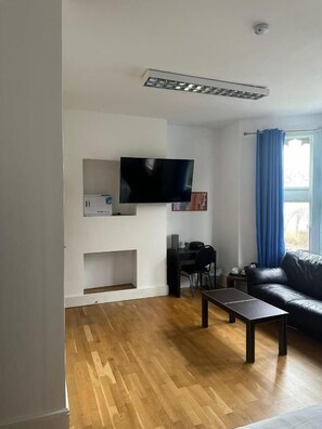 A living room with a flat-screen TV mounted on the wall, a black leather sofa, a wooden coffee table, and a blue curtain on the window. - 3 Luxury En-suite Bedrooms (Cardiff)