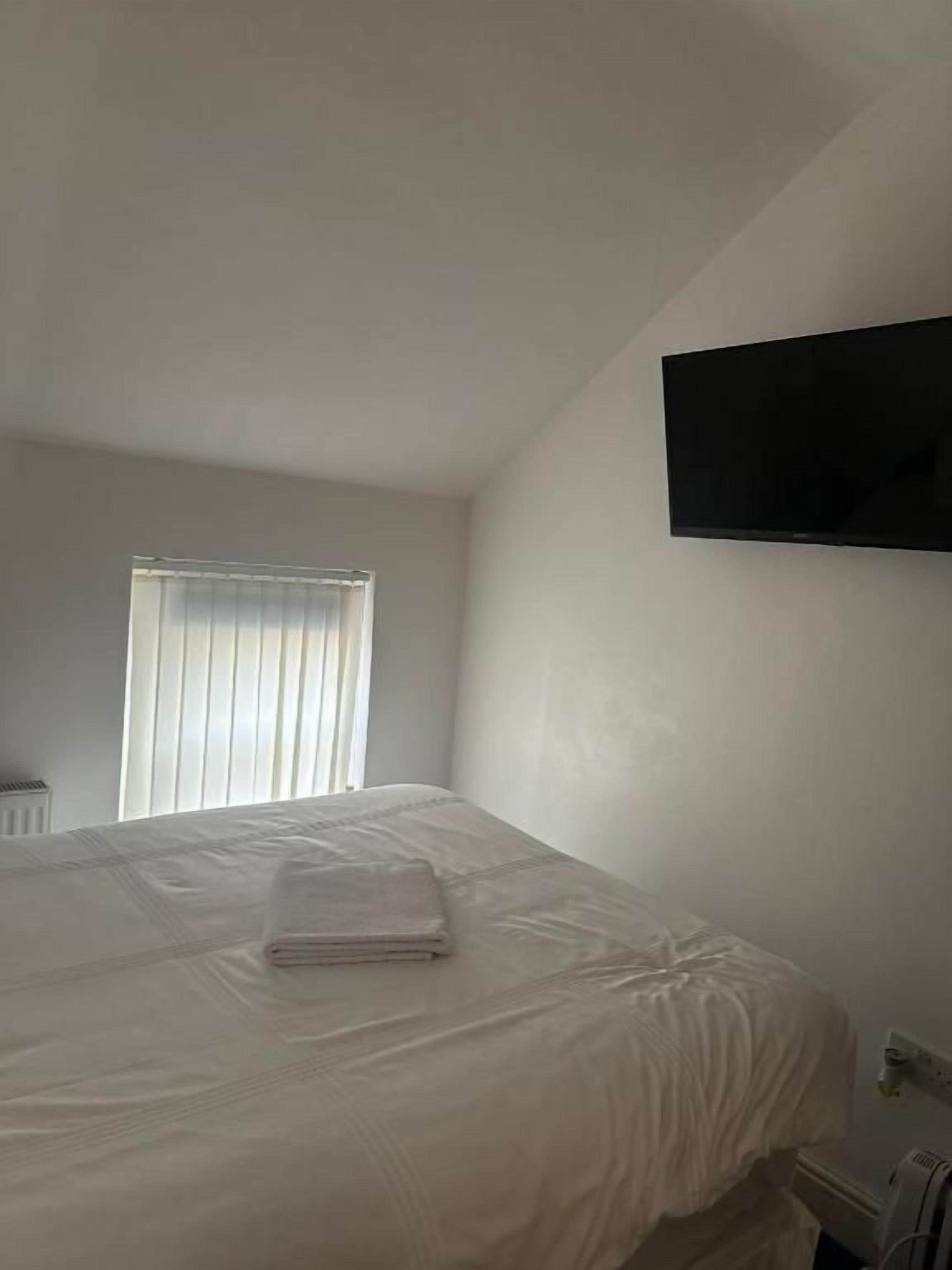 A hotel room with a bed, a flat-screen TV mounted on the wall, and a window with blinds.