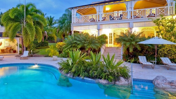 Ein Poolbereich mit Liegestühlen, ein zweistöckiges Gebäude mit Balkon und tropische Bepflanzung.