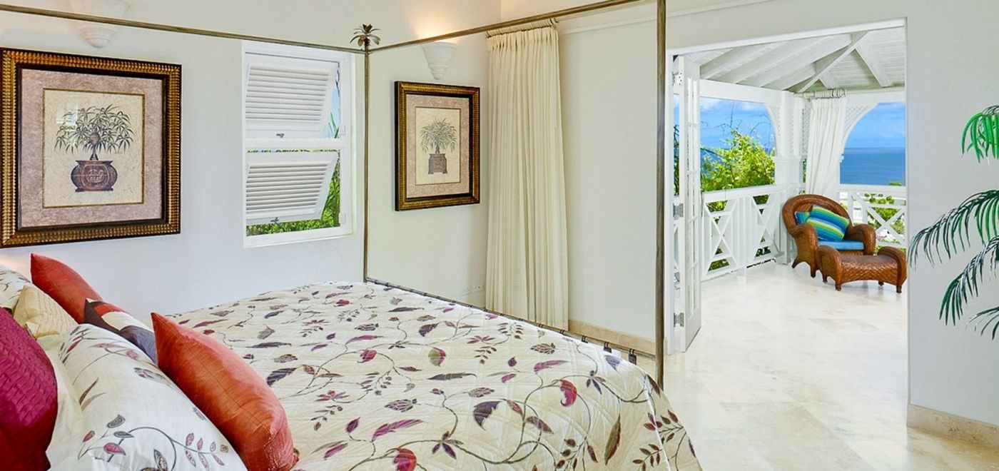 A bedroom with a bed, two framed pictures, a window with shutters, and a view of a balcony with a chair and a table.