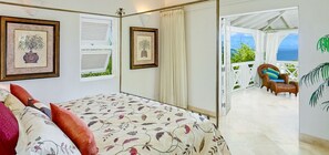 A bedroom with a bed, two framed pictures, a window with shutters, and a view of a balcony with a chair and a table. - Villa Calliaqua (Holetown)