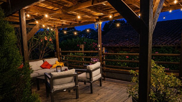 Una terraza de madera con un sofá y sillas, rodeada por una valla de madera y luces colgantes.