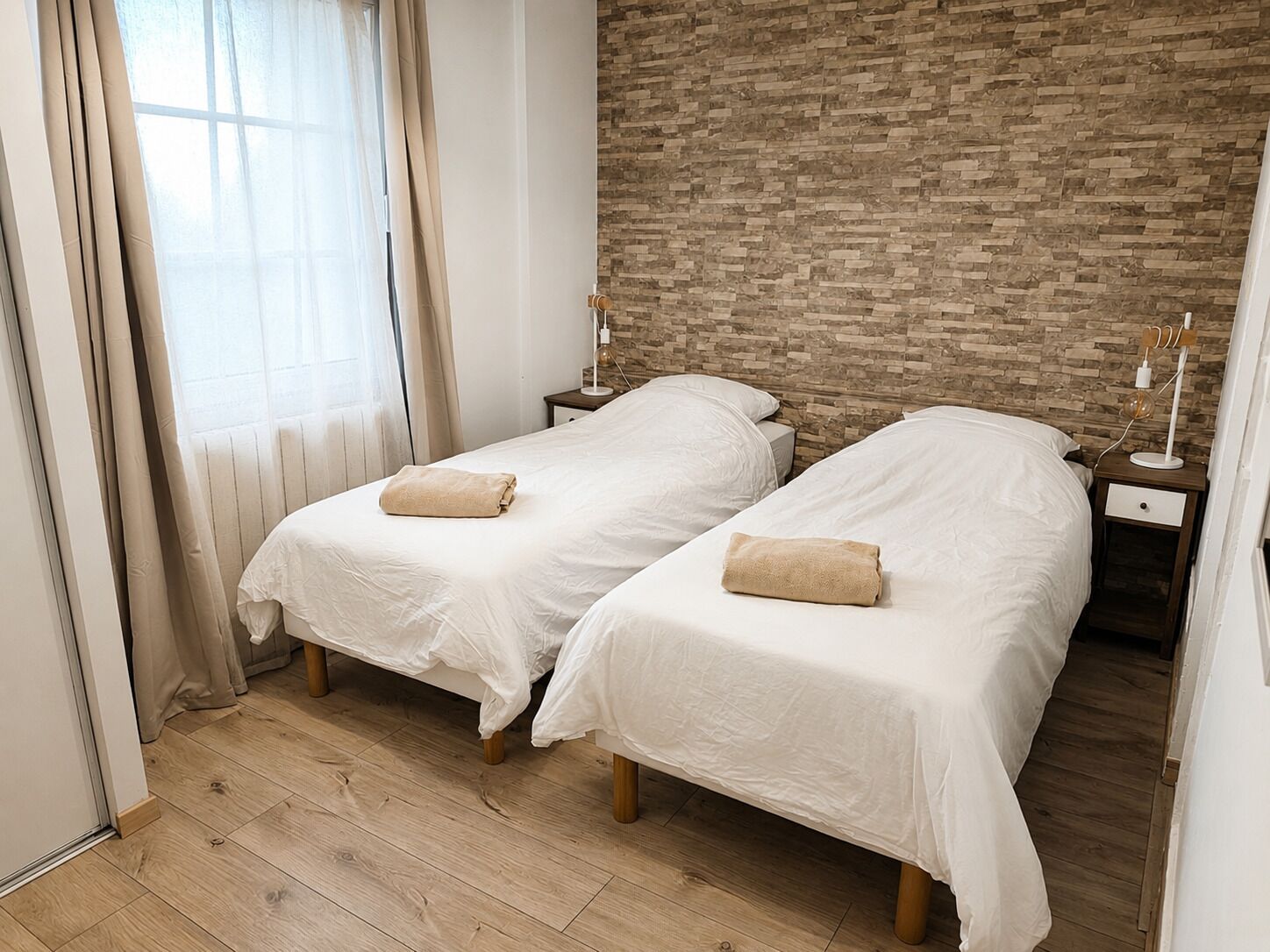 A hotel room with two single beds, a brick wall, a wooden floor, and a window with curtains.